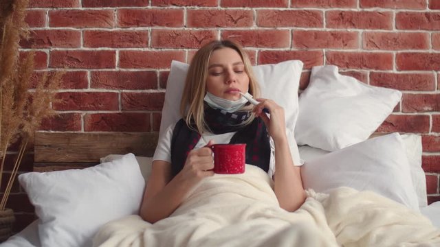 A young woman lies in bed covered with a blanket, takes a thermometer out of her mouth, takes a deep breath, and puts on a medical mask. A girl with a scarf around her neck and a mug of warm drink in