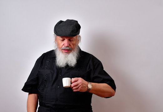 Old man enjoying his cup of expresso..Happy old man enjoys his coffee.. .Mature gentleman with a newsboy cap and black guayabera shirt and long white beard..