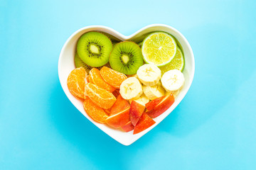Heart-shaped bowl with fruit on pastel blue background. The concept of healthy eating, eating fruit. Bowl with apple, mandarin banana. A bowl filled with fruit. Fruit and vitamins for the heart.