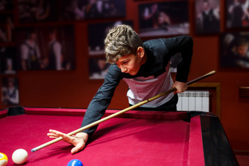Boy plays billiard or pool in club. Young Kid learns to play snooker. Boy with billiard cue strikes the ball on table. Active Leisure, sport, hobby concept	 - obrazy, fototapety, plakaty