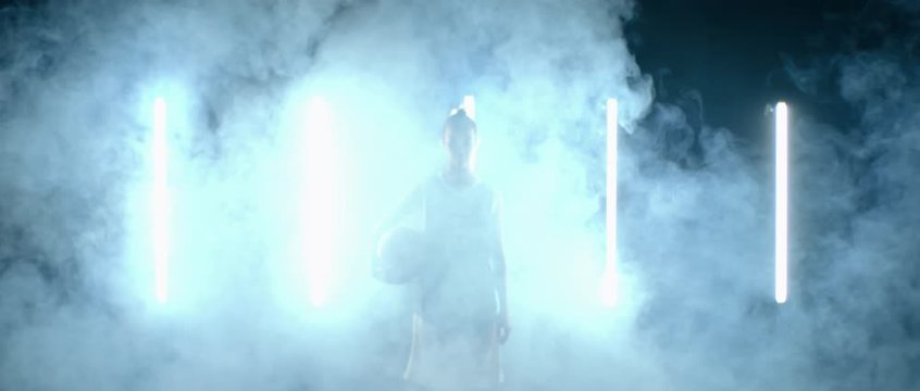 Caucasian female basketball player walking through the fog against bright lights, holding ball in her hands. 4K UHD 60 FPS SLOW MOTION shot on ARRI Alexa Mini with Atlas Orion 2x Anamorphic lens