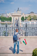 Enjoying vacation in Budapest. Young traveling man with national hungarian flag walking on riverside promenade with city view.