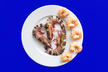 Fresh coocked boiled grilled and uncooked raw exotic black tiger big shrimps on white round plate on blue background, many king prawns. Healthy protein sea food. Top view.