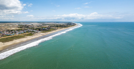 Aerial drone view of the beach, tourists, sea and waves splashing in the coast of the town Playas General Villamil, Ecuador. Sunny day. Beach houses and the horizon with some clouds in background.
