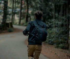 Teenager walking in the forest on wooden path with bag looking around. Adventuring wearing black jacket and beige pants.