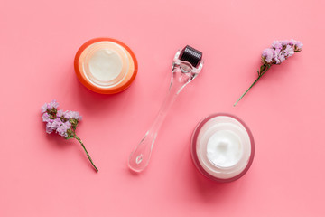 Cosmetologist tools. Dermaroller, creams near leaves and flowers on pink background top-down flat...