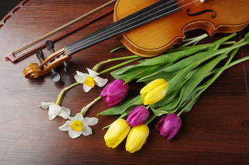 Violin with a bow, a bouquet of daffodils and tulips on a wooden table.