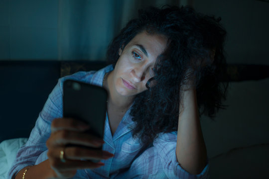 young tired and thoughtful hipster woman on bed late at night using social media app on mobile phone online dating in online addiction and internet overuse and obsession