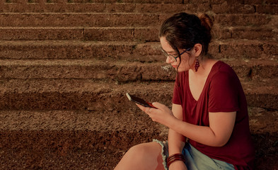 Beautiful Young girl of Indian ethnicity in her early 20s, wearing trendy casuals, is deeply engaged in her smartphone and accessing the internet for social media status update using public wifi.