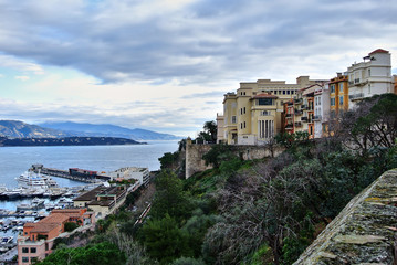 Fototapeta na wymiar Monte Carlo, Monaco - Houses in bright colors on the shores of the Mediterranean Sea, a bay with moorings on which there are yachts, the sky with white-gray clouds.