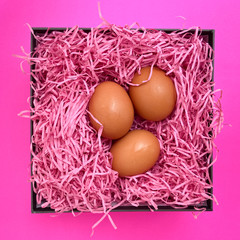 Three chicken brown eggs in the pink hay inside a square box is place on pink background. Close up, center focus for easter, agriculture, poultry and cooking theme.