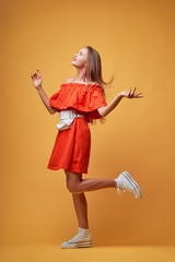 Summer urban fashion. Fun and colorful. Young pretty happy woman in orange dress posing against yellow background.