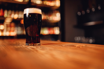 Glass of dark beer craft with foam on wooden bar counter, pub blurred background