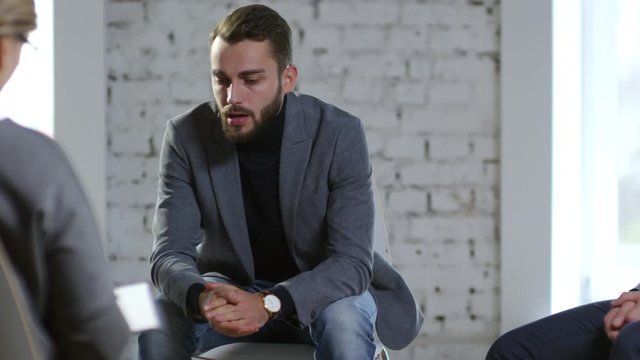 PAN of rich businessman with stylish beard talking about his addiction problems to psychotherapist and patients during interpersonal group therapy