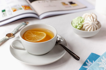 White cup of green tea with lemon, mini meringue in a bowl on a white bedsheet with an illustrated magazine, watch and tea bags. Optimistic lifestyle