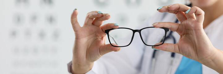 Focus on female oculist standing in clinic office and holding new glasses. Smiling ophthalmologist wearing white medical uniform. Vision test and healthcare concept