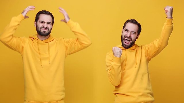 Two bearded young guys 20s in hoodie isolated on yellow background studio. People lifestyle concept. Looking at camera Doing winner gesture say Yes, sad upset cry expressive gesticulating with hands