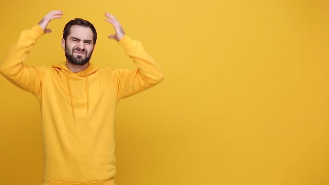 Handsome bearded young guy 20s in streetwear hoodie isolated on yellow background studio. People sincere emotions lifestyle concept. Looking at camera sad upset cry expressive gesticulating with hands