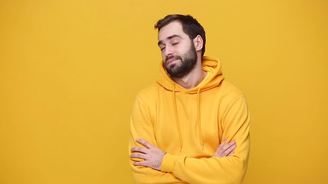 Handsome sad exhausted boring bearded young guy 20s in streetwear hoodie isolated on yellow background studio. People sincere emotions, lifestyle concept. Looking at camera with folded crossed hands