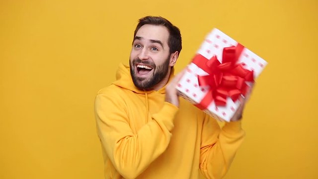 Bearded young guy 20s in streetwear hoodie isolated on yellow background studio. People lifestyle concept. Looking at camera, charming smile, hold white red polkadot present box with gift ribbon bow