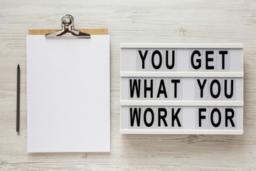 'You get what you work for' words on a lightbox, clipboard with blank sheet of paper on a white wooden background, overhead view. Top view, from above, flat lay.