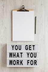 'You get what you work for' words on a lightbox, clipboard with blank sheet of paper on a white wooden background, overhead view. Top view, from above, flat lay.