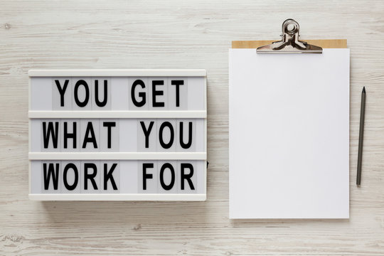 'You get what you work for' words on a modern board, clipboard with blank sheet of paper on a white wooden background, overhead view. Top view, from above, flat lay.