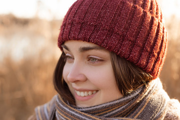 Close up portrait of a pretty smiling young girl in a park. Lady warms up in a hat and scarf. Woman in nature enjoys the weather.