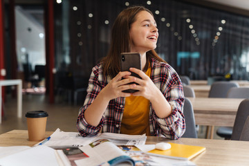 Image of joyful beautiful girl using cellphone while doing homework