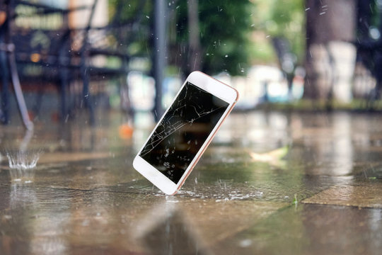  White smartphone falling and crashing on wet ground in the city park on a rainy day