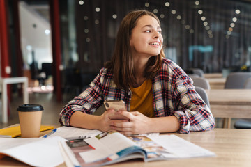 Image of joyful caucasian girl using cellphone while doing homework