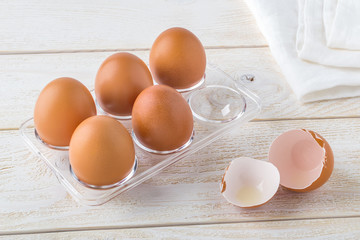 Five raw chicken eggs in a transparent plastic tray and empty eggshell near it on a white wooden table. Cooking for easter with brown eggs.