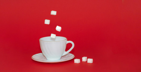 Levitation of sugar cubes over a white mug with a saucer on a red isolated background. Levitation of objects.Minimalism or surrealism. The concept of the sweet life. Banner, copy space.