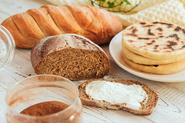  Loaf of rye bread and a sandwich with butter on a background of bread and tortillas. Baking for breakfast on a white wooden table.