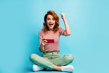 Full length photo funky impressed woman sit floor legs crossed use smartphone win online social network lottery raise fists scream wow wear green stylish jumper isolated blue color background