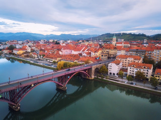 Fototapeta na wymiar Aerial view of Maribor city in Slovenia on the banks of Drava river. Scenic landscape with buildings, embankment, historical bridge, mountains and blue cloudy sky, travel background, Lower Styria