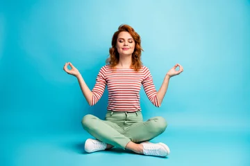 Fototapeten Foto in voller Größe einer ruhigen, ruhigen Frau, die auf dem Boden gekreuzte Beine sitzt, zeigt ein Om-Zeichen, eine Yoga-Meditation, die ein stilvolles grünes Outfit einzeln auf blauem Hintergrund trägt © deagreez