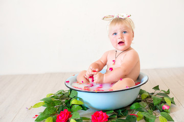 Little cute baby girl is sitting in a basin with pink petals and strawberries.