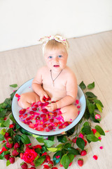 Little cute baby girl is sitting in a basin with pink petals and strawberries.