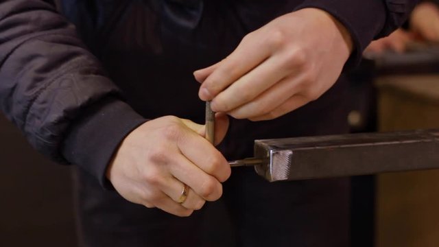 Static close up of a metalworker using a t-shaped threading tool to cut a hole into the end of a metal bar with other workers in the background