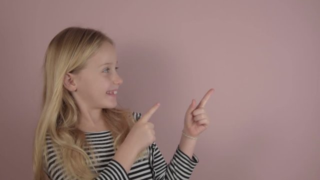 pretty smiling blonde little girl points to something posing on light pink background in light studio closeup