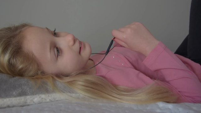 pretty blonde girl in pink sweater talks on phone with headset lying on soft bed in light bedroom at home closeup