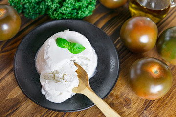 A fresh soft ricotta cheese with basil leaf on black plate, wooden tables with tomatoes, olive oil,...