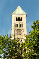 Mächtiger Glockenturm mit großem Ziffernblatt und Marienstatue der Heilig-Geist-Kirche, eine neuromanische Basilika in der Stadt Schweinfurt bei strahlend blauem Himmel, im Vordergrund grüne Bäume