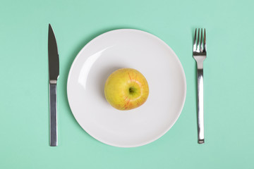 Whole green apple on a white plate with fork and knife lying near, top view.
