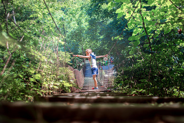 A girl walks along a rickety wooden rope bridge among a green forest above a mountain stream.