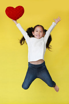 Smiling Asian Little Girl Jumping On Air With Holding Red Heart In Hand Isolated On Yellow Background.