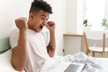 Happy positive african guy using laptop computer in bed.