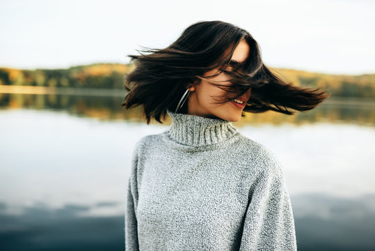 Image of happy young brunette woman with closed eyes, wearing grey sweater, posing on nature background. Pretty girl posing against the lake in the park.