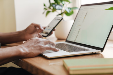 Guy indoors using laptop computer and mobile phone.